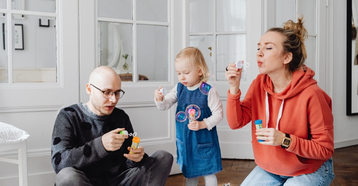 Twee ouders en een jong kind spelen samen met zeepbellen in huis. Het gezin geniet zichtbaar van een vrolijk en ontspannen moment samen.