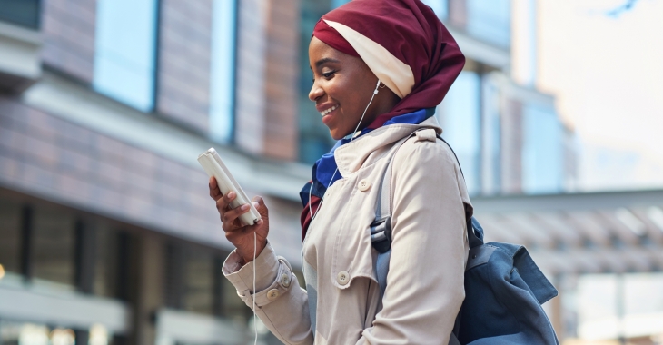 Vrouw met hoofddoek kijkt al lachend naar smartphone en luistert naar muziek via oortjes