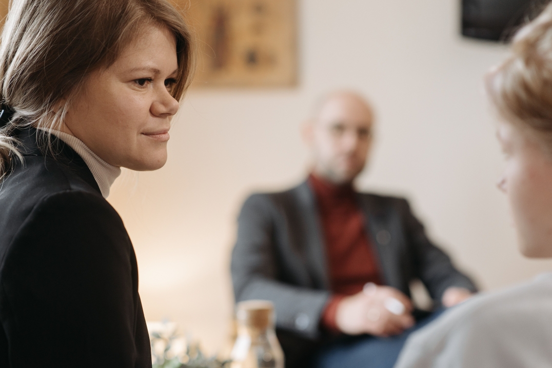 Begeleider luistert aandachtig tijdens een gesprek met een cliënt, terwijl een collega op de achtergrond aanwezig is.