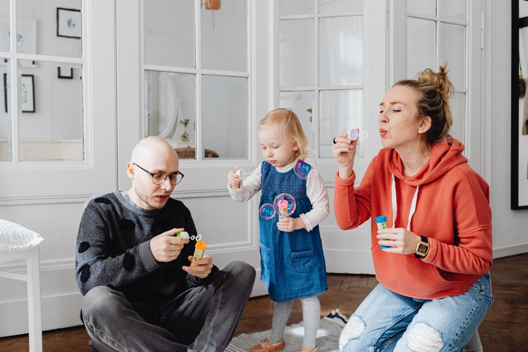 Twee ouders en een jong kind spelen samen met zeepbellen in huis. Het gezin geniet zichtbaar van een vrolijk en ontspannen moment samen.
