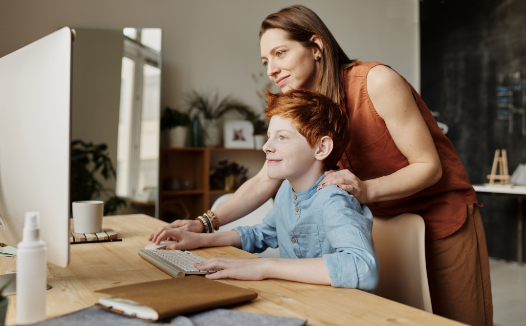 Een ouder helpt een kind achter een computer terwijl ze samen aandachtig naar het scherm kijken aan een bureau thuis.