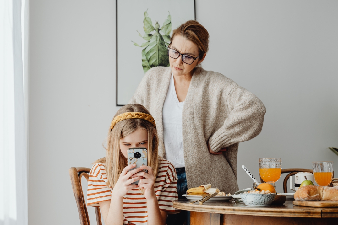 Kind kijkt naar een smartphone aan de ontbijttafel terwijl een ouder bezorgd toekijkt.