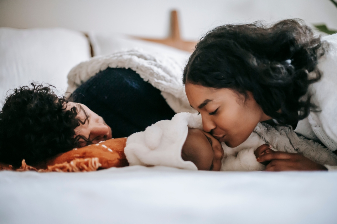 Twee ouders liggen op bed naast hun slapende baby en geven het kind liefdevol een kus. De warme en intieme scène straalt geborgenheid, zorg en familieband uit.