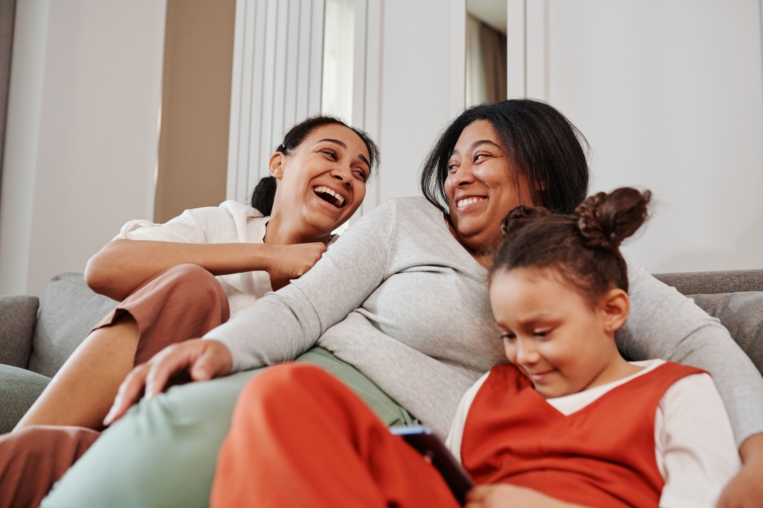 Twee vrouwen en een kind lachen samen op de bank in een huiselijke omgeving.