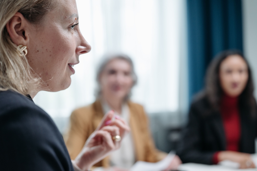 Persoon spreekt tijdens een overleg terwijl andere deelnemers aandachtig luisteren.